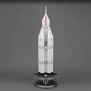 May include: A white and red model of a Soviet-era rocket, the R-7, on a silver stand. The rocket has four booster rockets and a pointed nose cone. The rocket is labeled "CCCP" on the side.