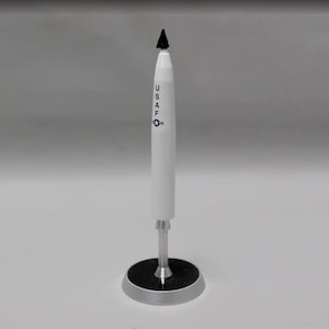 Może przedstawiać: Biały model rakiety z czarnym, spiczastym stożkiem nosowym. Rakieta ma napis "USAF" nadrukowany na boku. Rakieta stoi na czarnej i srebrnej podstawie.