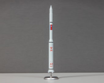 Pode incluir: Um modelo de foguete branco com uma bandeira vermelha e azul no lado. O foguete tem as palavras coreanas "새" e "로" escritas nele.