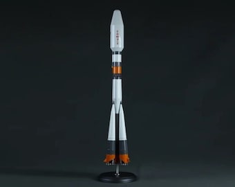 Pode incluir: Um modelo de foguete branco e laranja com a palavra "Soyuz" escrita no lado. O foguete está em um suporte preto com três pernas.