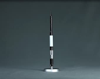 Pode incluir: Um modelo de foguete preto e branco com uma base prateada. O foguete tem um topo pontiagudo e um corpo cilíndrico com seções brancas e pretas. Ele se ergue sobre um fundo cinza.