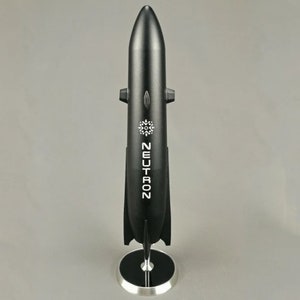 May include: A black model rocket with the word "NEUTRON" printed on the side. The rocket is on a silver stand with a black base.