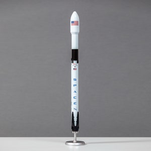 Puede incluir: Un modelo de cohete blanco y negro con las palabras "SpaceX" y "Falcon 9" impresas en el lateral. El cohete tiene una bandera estadounidense en la parte superior y está sobre un soporte negro.