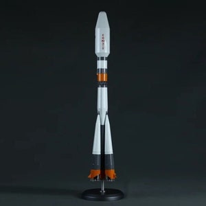 May include: A white and orange model rocket with the word "Soyuz" written on the side. The rocket is on a black stand with three legs.