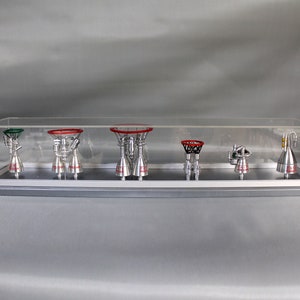 以下が含まれることがあります： 7つの銀色のロケットエンジンのモデルが入ったディスプレイケース。エンジンは赤と緑のアクセントが付けられており、灰色のベースに一列に並んでいます。