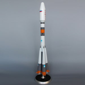 May include: A white and orange model rocket with the word "Soyuz" written on the side. The rocket is on a black stand with a silver base.