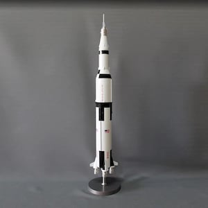 May include: A white and black model of the Saturn V rocket, a large, multi-stage rocket used in the Apollo program. The model is on a silver stand and has a US flag on the side.