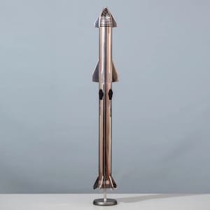 May include: A silver model of a rocket ship on a stand. The rocket has a pointed nose cone and a cylindrical body with fins at the base.