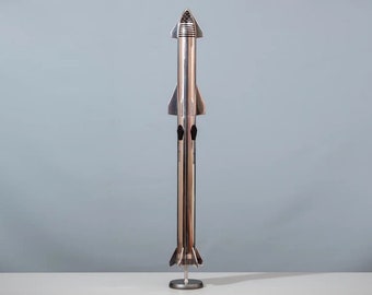 Pode incluir: Um modelo prateado de um foguete em um suporte. O foguete tem um cone de nariz pontudo e um corpo cilíndrico com aletas na base.