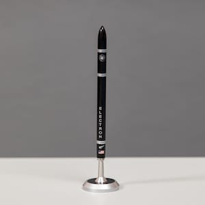 Könnte beinhalten: Ein schwarzer Stift in Form einer Rakete mit dem Wort "ELECTRON" auf der Seite. Der Stift steht auf einem silbernen Ständer.