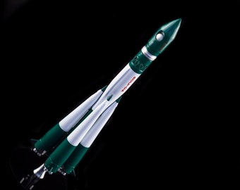 Pode incluir: Um modelo de foguete verde e branco com a palavra "ВОСТОК" em letras vermelhas no lado. O foguete está sobre um fundo preto.