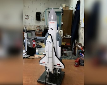 Pode incluir: Um modelo branco e cinza da nave espacial soviética Buran, montado em um suporte preto. A nave espacial é etiquetada como "CCCP" e "ЭНЕРГИЯ".