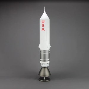 May include: A white model rocket with a red "USA" inscription on the side. The rocket is on a gray stand with a round base.