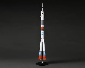 Pode incluir: Um modelo de foguete branco e cinza com detalhes laranja e uma base preta. O foguete tem um logotipo azul e branco no lado.