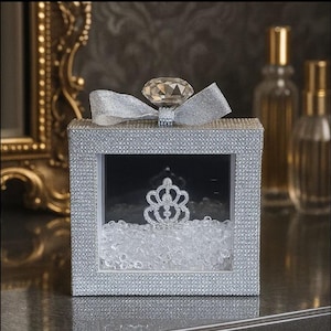 Könnte beinhalten: Eine silberne, quadratische Dekorationsbox, besetzt mit Strasssteinen. Sie hat ein klares Fenster, das eine silberne Krone auf klaren Perlen zeigt. Ein großer, facettierter Kristall sitzt oben auf der Box, verziert mit einem silbernen Band.