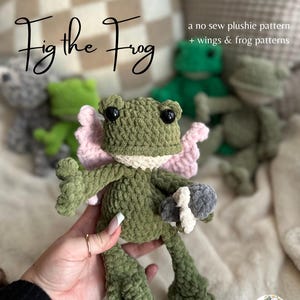 Puede incluir: Una rana de peluche verde hecha a mano con alas rosas y un cuello blanco. La rana tiene ojos negros y sostiene un objeto gris y blanco. El texto "Fig the Frog" está encima de la rana, con texto adicional.