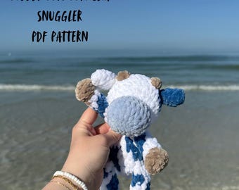May include: A handmade, plush cow toy with white, blue, and brown accents. The cow is held in front of a beach scene. Text on the image reads "CUDDLY COW CORNELIA SNUGGLER PDF PATTERN" and "BP: Snugglers".