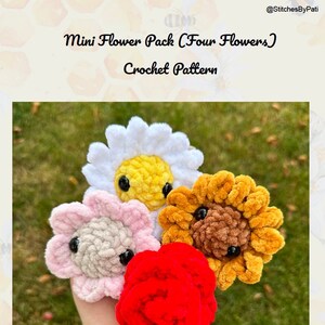 May include: Four crocheted flower plushies in different colours: white, pink, brown, and red. The flowers are arranged in a hand, with the white flower at the back and the red flower at the front. The flowers have black eyes and are made with fuzzy yarn.