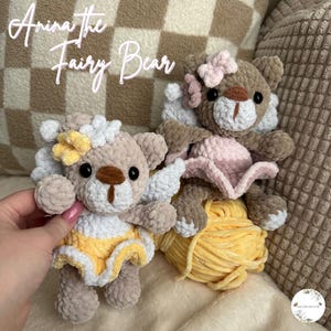 May include: Two crocheted teddy bears dressed as fairies. The bear on the left is wearing a yellow dress with white accents and a white flower crown. The bear on the right is wearing a pink dress with white accents and a pink flower crown. Both bears have brown fur and are sitting on a yellow yarn ball.