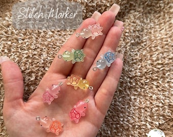 Puede incluir: Una colección de marcadores de punto de tortuga coloridos. Los marcadores son de varios tonos de rosa, naranja, amarillo, verde y azul. Cada tortuga tiene un lazo plateado para sujetar a los proyectos de tejido. El texto "Turtle Stitch Marker" está en la esquina superior izquierda.