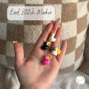 May include: A hand displays five bird stitch markers. These small, colourful bird charms include a pink flamingo, a black toucan, and white birds with yellow accents. The text "Bird Stitch Marker" is in a white rectangle.