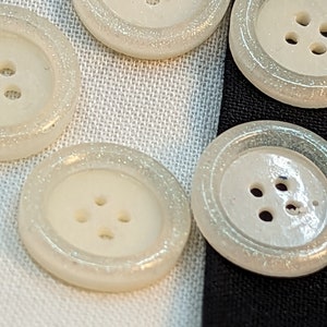 Könnte beinhalten: Fünf weiße Knöpfe mit glitzernder Oberfläche und jeweils vier Löchern. Die Knöpfe sind in einem Halbkreis auf einem weißen und schwarzen Stoffhintergrund angeordnet.