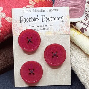 Op de afbeelding: Drie rode glitterharsknoppen met zwarte stiksels. De knoppen worden getoond op een kaart met de tekst "From Metallic Visions: Bobbie's Buttonry Hand-made unique resin buttons".