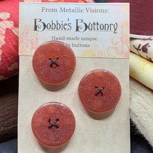 Op de afbeelding: Drie rode glitterknopen met zwarte stiksels. De knopen worden getoond op een kaart met de tekst "From Metallic Visions: Bobbies' Buttonry Hand-made unique buttons".