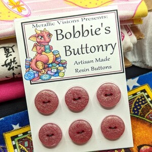 Op de afbeelding: Zes roze glitterharsknoppen met zwarte stiksels. De knoppen worden getoond op een witte kaart met de tekst "Metallic Visions Presents: Bobbie's Buttonry Artisan Made Resin Buttons".