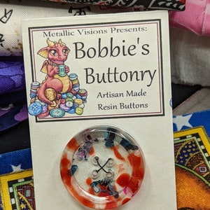 May include: A red, white, and blue resin button with a clear background. The button is surrounded by a white card with the text "Metallic Visions Presents: Bobbie's Buttonry Artisan Made Resin Buttons".
