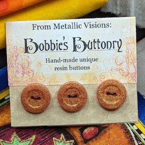 Op de afbeelding: Drie ronde, oranje, glimmende harsknoppen met een zwart midden. De knoppen bevinden zich op een kaart met de tekst "From Metallic Visions: Bobbie's Buttonry Hand-made unique resin buttons".