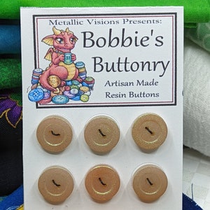 Könnte beinhalten: Sechs irisierende goldene Harz-Knöpfe mit jeweils zwei Löchern, auf einer weißen Karte mit dem Text "Metallic Visions Presents: Bobbie's Buttonry Artisan Made Resin Buttons" dargestellt.