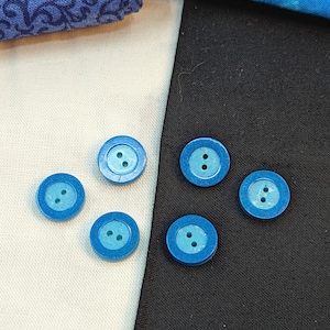 Op de afbeelding: Zes blauwe knopen met een lichtblauw midden en twee gaten. De knopen zijn in een halve cirkel op een witte en zwarte stoffen achtergrond gerangschikt.