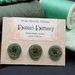 Op de afbeelding: Drie groene harsknoppen met gouden draadaccenten. De knoppen worden gepresenteerd op een kaart met de tekst "From Metallic Visions: Bobbie's Buttonry Hand-made unique resin buttons".