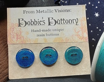 May include: Three blue and green resin buttons with a black line across the center. The buttons are on a card that says "From Metallic Visions: Bobbie's Buttonry Hand-made unique resin buttons".