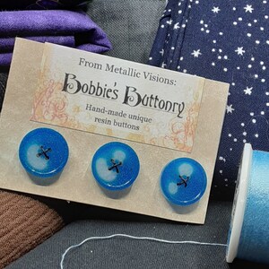 Op de afbeelding: Drie blauwe harsknoppen met een wit swirl-ontwerp en een zwart kruis in het midden. De knoppen worden getoond op een kaart met de tekst "From Metallic Visions: Bobbie's Buttonry Hand-made unique resin buttons".