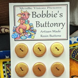 Könnte beinhalten: Sechs goldfarbene Glitzer-Harzknöpfe auf einer weißen Karte mit dem Text "Metallic Visions Presents: Bobbie's Buttonry Artisan Made Resin Buttons".