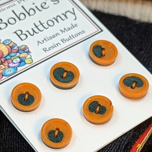 Op de afbeelding: Zes oranjeharsknoppen met zwarte centra. De knoppen zijn in een cirkel gerangschikt op een witte kaart met de tekst "Bobbie's Buttonry Artisan Made Resin Buttons" erop gedrukt.