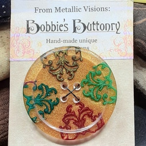 Op de afbeelding: Een ronde, gouden knoop met een glinsterende afwerking. De knoop heeft vier gaten en is versierd met groene, rode en gouden bloemmotieven. De tekst "Bobbie's Buttonry" en "Hand-made unique" is gedrukt op een kaart achter de knoop.