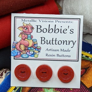 Op de afbeelding: Drie rode glitterharsknoppen met een zwart lijntjesontwerp. De knoppen bevinden zich op een witte kaart met de tekst "Metallic Visions Presents: Bobbie's Buttonry Artisan Made Resin Buttons".
