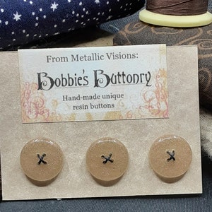 Op de afbeelding: Drie bruine harsknoppen met een zwart X-ontwerp. De knoppen worden weergegeven op een kaart met de tekst "From Metallic Visions: Bobbie's Buttonry Hand-made unique resin buttons".