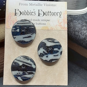 Op de afbeelding: Drie handgemaakte unieke knopen met een abstract ontwerp in zwart en wit. De knopen bevinden zich op een kaart met de tekst "From Metallic Visions: Bobbie's Buttonry Hand-made unique buttons".
