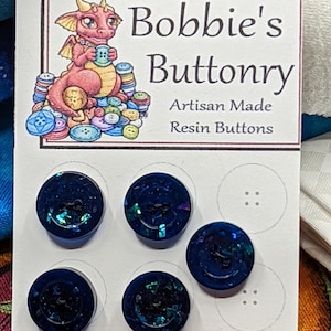 May include: Five blue resin buttons with iridescent glitter embedded in the resin. The buttons are displayed on a white card with the text "Metallic Visions Presents: Bobbie's Buttonry Artisan Made Resin Buttons".