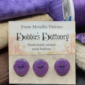 Op de afbeelding: Drie paarse harsknoppen met een glinsterende afwerking en zwarte centra. De knoppen worden getoond op een kaart met de tekst "From Metallic Visions: Bobbies' Buttonry Hand-made unique resin buttons."