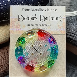 Op de afbeelding: Een regenboogkleurige harsknop met een helder midden en een kruisvormig ontwerp. De knop bevindt zich op een kaart met de tekst "From Metallic Visions: Bobbie's Buttonry Hand-made unique resin buttons".