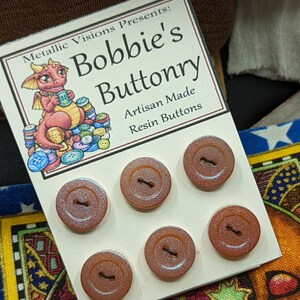 Op de afbeelding: Zes iriserende bruine harsknoppen met een glinsterende afwerking. De knoppen worden gepresenteerd op een witte kaart met de tekst "Metallic Visions Presents: Bobbie's Buttonry Artisan Made Resin Buttons".