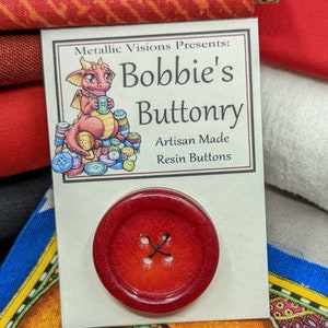 Op de afbeelding: Een rode harsknop met een ontwerp met vier gaten. De knop bevindt zich op een kaart met de tekst "Metallic Visions Presents: Bobbie's Buttonry Artisan Made Resin Buttons".