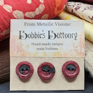 Op de afbeelding: Drie handgemaakte harsknoppen met een zwart en rood marmerpatroon. De knoppen worden gepresenteerd op een kaart met de tekst "From Metallic Visions: Bobbie's Buttonry Hand-made unique resin buttons".