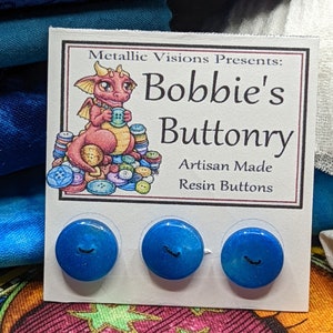 Op de afbeelding: Drie blauwe harsknoppen met een wit swirl-ontwerp en een zwarte smiley. De knoppen worden weergegeven op een witte kaart met de tekst "Metallic Visions Presents: Bobbie's Buttonry Artisan Made Resin Buttons".