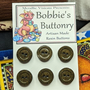 Op de afbeelding: Zes gouden glitterharsknoppen met elk twee gaten. De knoppen worden getoond op een witte kaart met de tekst "Metallic Visions Presents: Bobbie's Buttonry Artisan Made Resin Buttons".